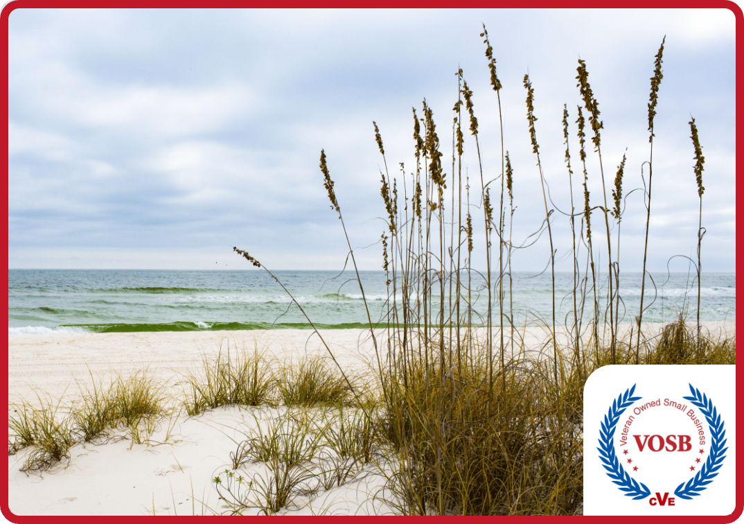 Beautiful beach with white sand and beach grass with teal water and cloudy blue skies in the distance.