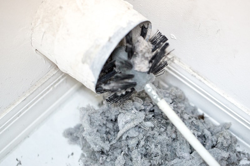 Dryer Vent Cleaning. Dryer vent in a home being cleaned out with a round brush. There is a large pile of lint that has been removed from the vent on a white tiled floor. The walls and baseboards are white. The lint is gray.